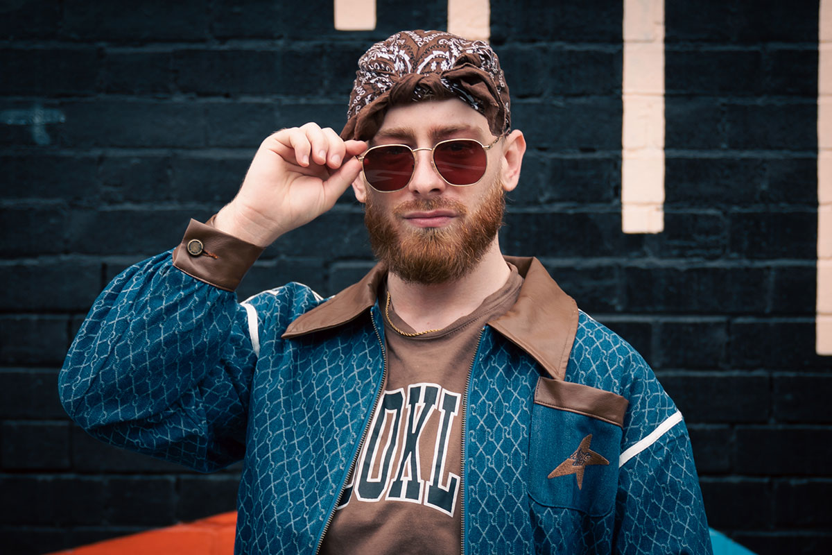 Headshot of student looking towards the camera wearing a blue and brown jacket, brown paisley bandana, and sunglasses which they are holding at the side.