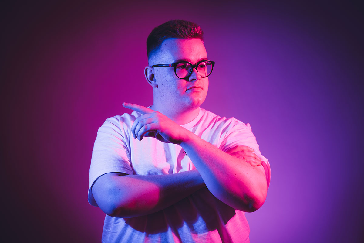 Headshot of student looking to their left with their arms crossed and one hand slightly raised to point in the opposite direction of viewing. They're lit by bright pink and purple lighting.