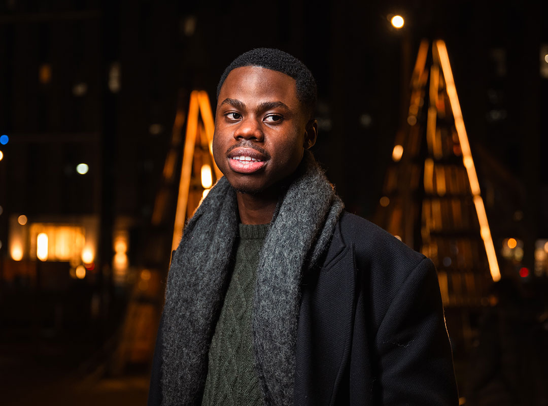 A student in a coat and scarf stands in front of an illuminated building at night.