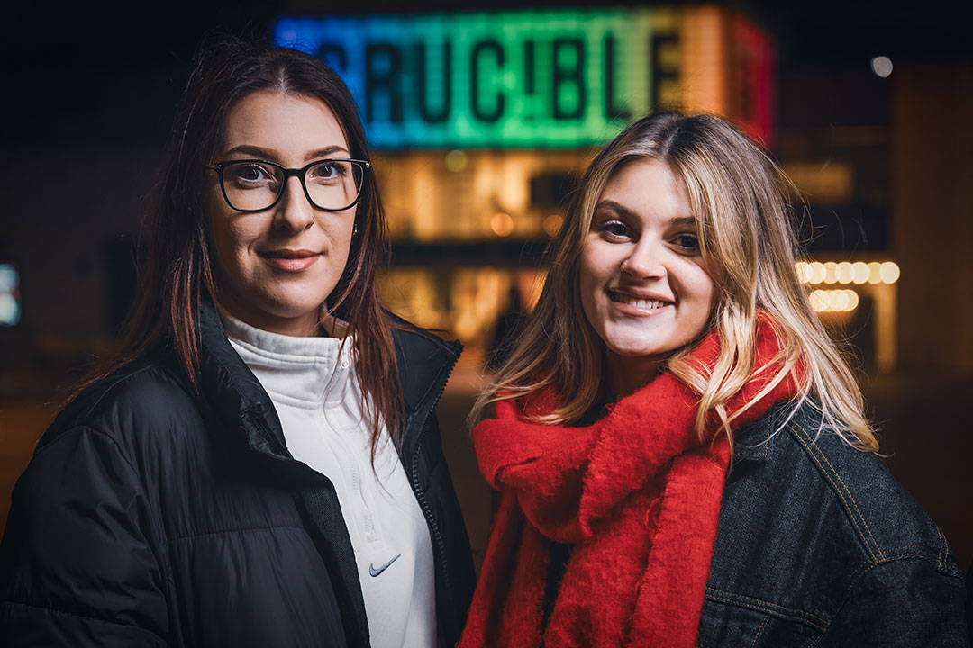 Two students stand smiling in front of the Crucible's bright, colourful sign. One wears glasses and a black jacket, the other a red scarf.