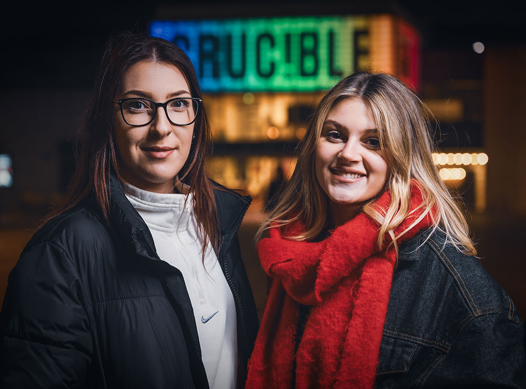 Two students stand smiling in front of the Crucible's bright, colourful sign. One wears glasses and a black jacket, the other a red scarf.