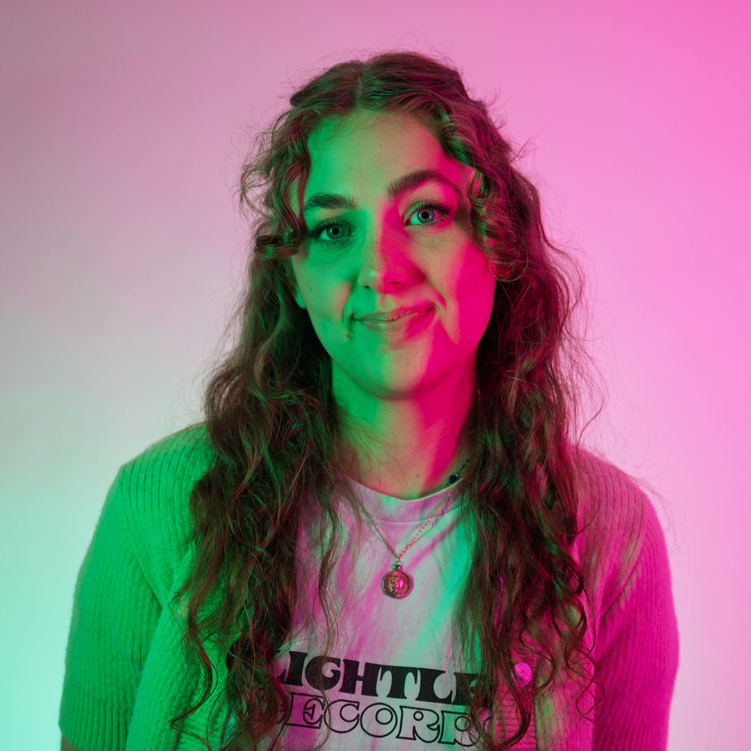 Molly Pemberton lit by bright pink lights, looking to the side of the camera smiling and resting her arm on her knee.