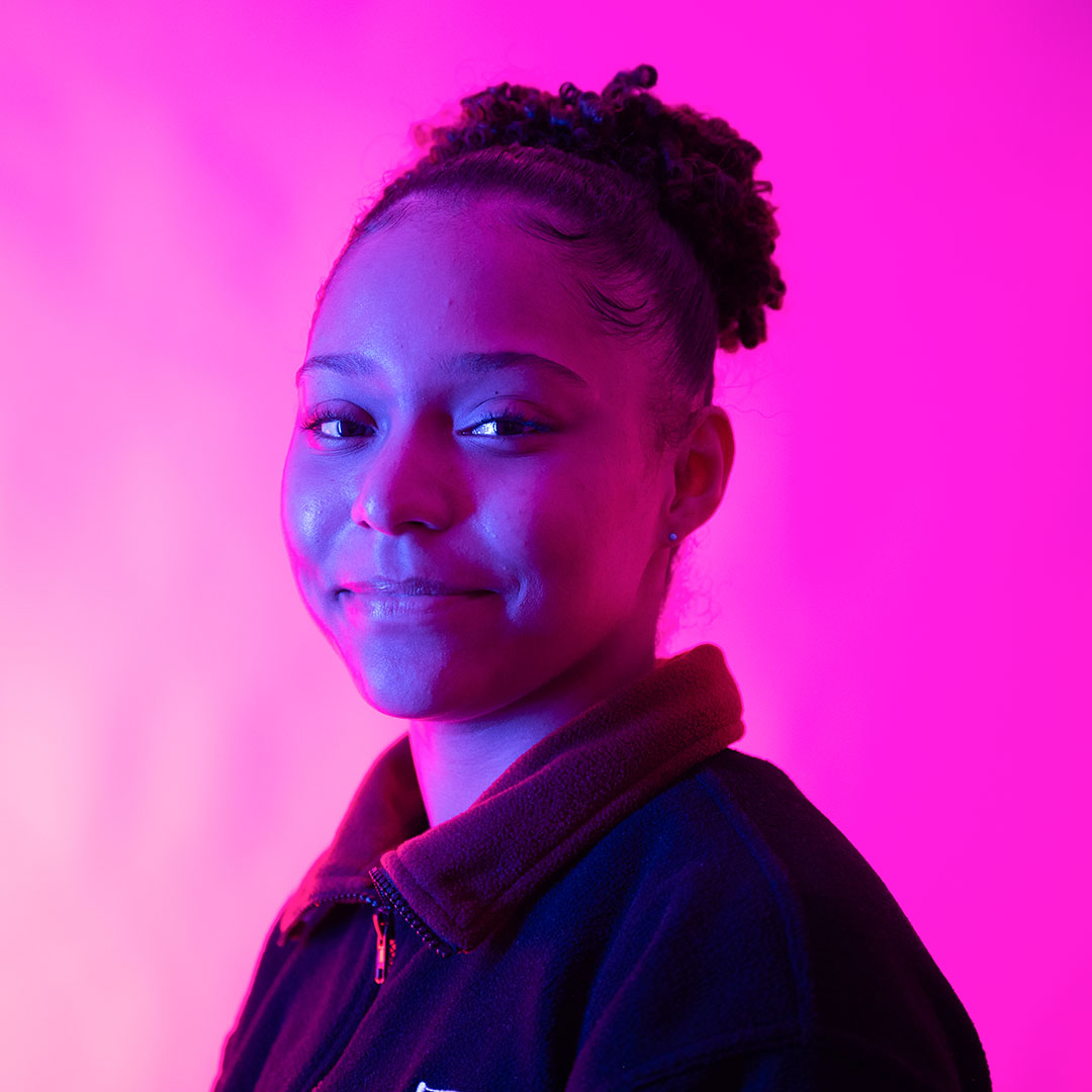 Layla Barrett lit by bright pink and blue lights, looking towards the camera smiling.