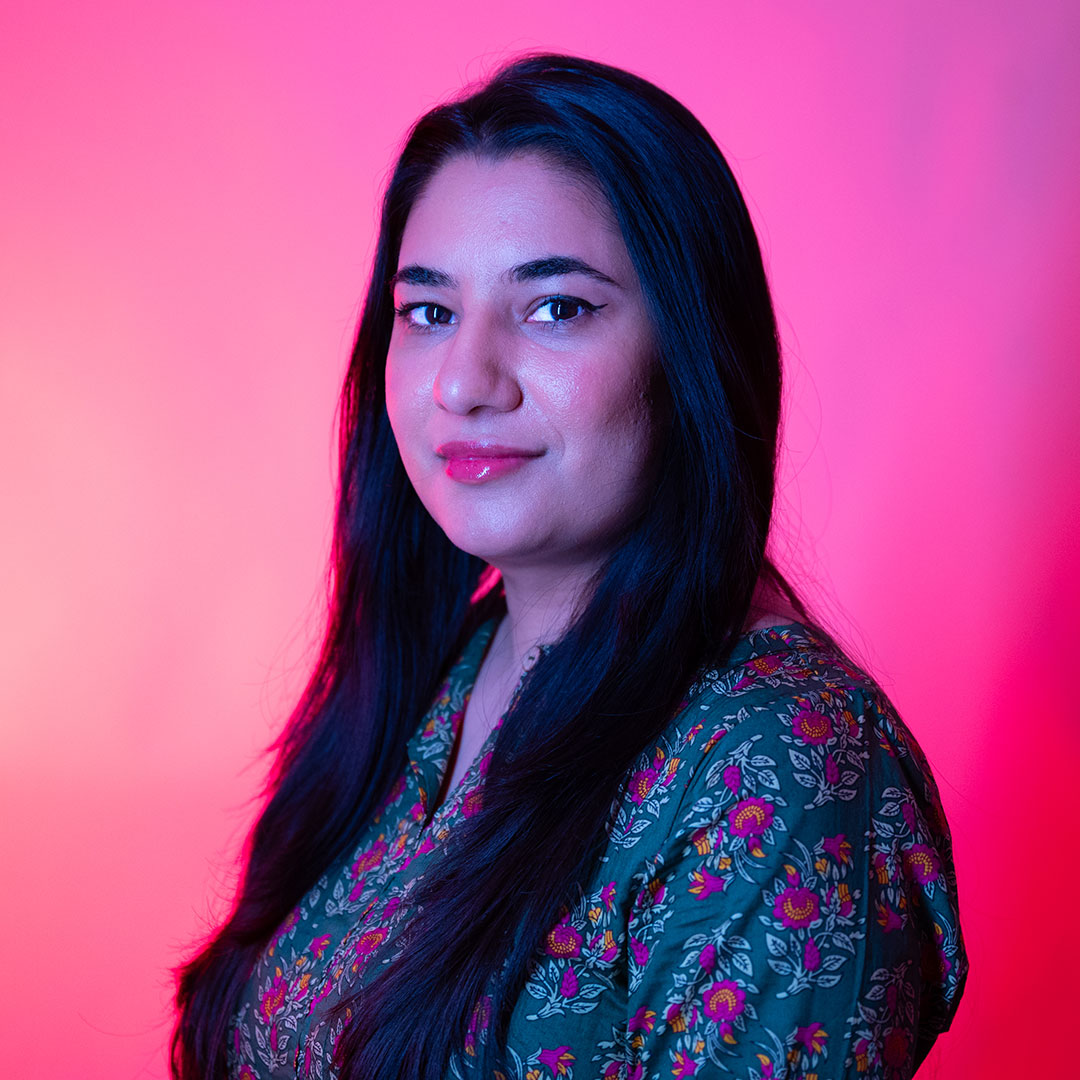 Shafaq Sajid lit by bright pink lights, looking towards the camera smiling.