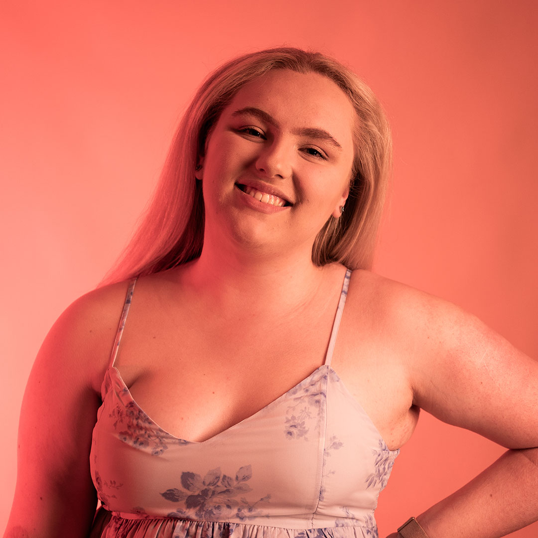 Abi Marchant lit by bright orange lights, looking towards the camera smiling with her hand on her hip.