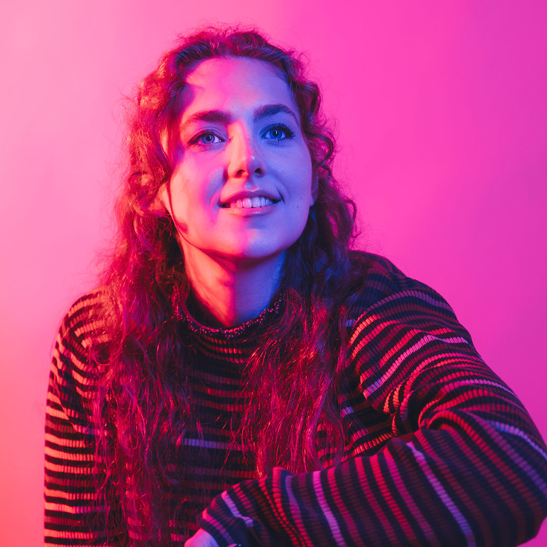 Molly Pemberton lit by bright pink lights, looking to the side of the camera smiling and resting her arm on her knee.
