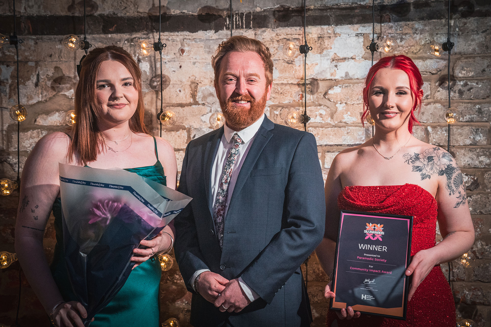 Three award winners pose happily in front of a brick wall celebrating their success. One holds a bouquet of flowers and another holds their framed award certificate.