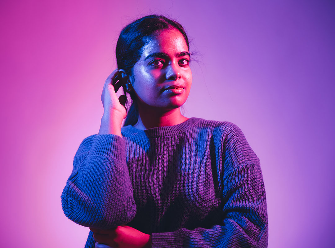 Student lit with bright purple light looking towards the camera with a neutral expression. One arm is folded across their front and the other is bent with their hand resting on their ear.