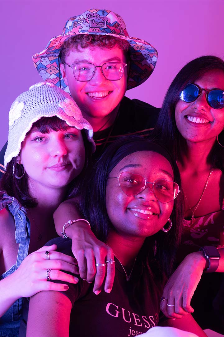 Four students smiling at the camera, there's pink and blue neon lighting around them