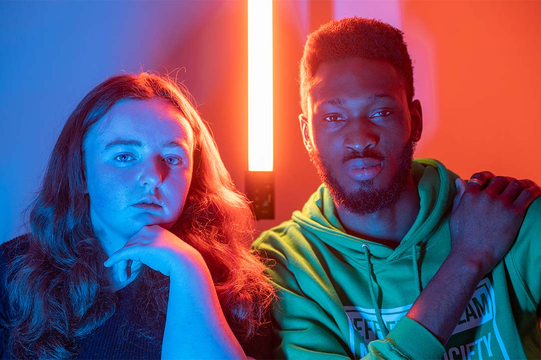 Two students smiling at the camera, there's pink and blue neon lighting around them